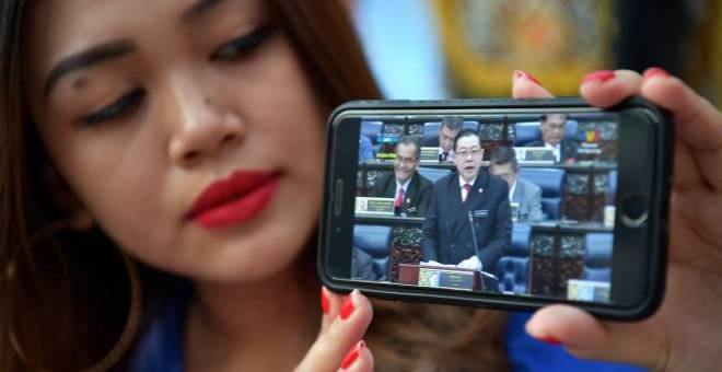 Maryanne Lee, 25, mengikuti siaran langsung pembentangan Belanjawan 2019 di Parlimen oleh Menteri Kewangan Lim Guan Engmelalui telefon bimbit di Tebingan Sungai Kuching, hari ini.