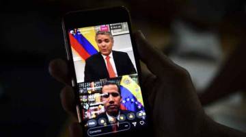 KECAM AS: Seorang wartawan mengikuti perbualan secara langsung dalam talian antara Presiden Colombia Ivan Duque (atas) dan Guaido menerusi telefon pintar di Caracas kelmarin. Maduro mengecam AS kelmarin kerana ‘mencuri’ berbilion dolar dan memberi ‘sisa’ untuk bantuan kemanusiaan. — Gambar AFP